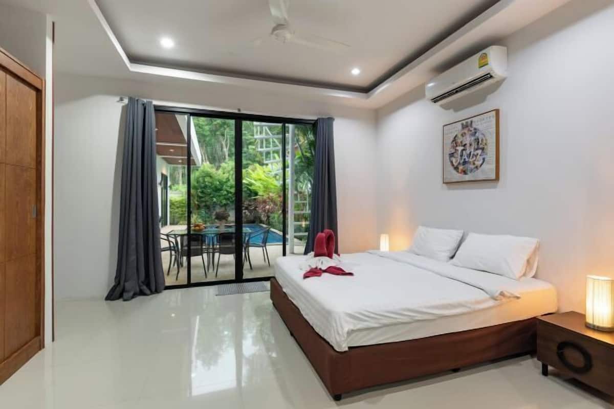 Une chambre à coucher comprenant un lit, une table de chevet, un ventilateur de plafond, un appareil de climatisation, une porte-fenêtre coulissante donnant sur un balcon et un tableau encadré accroché au mur.