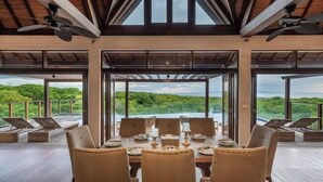 A dining area with a table set for six, large windows offering a view of a lush landscape, and ceiling fans. - Khaya Luxury Villas (Bali)