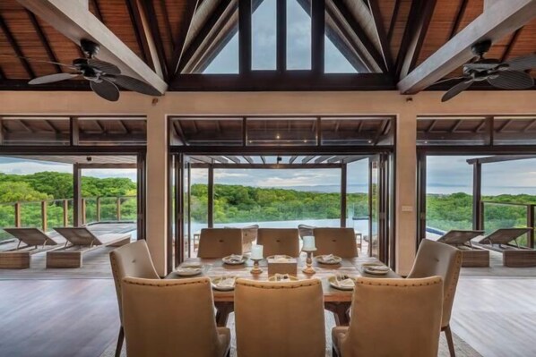 A dining area with a table set for six, large windows offering a view of a lush landscape, and ceiling fans. - Khaya Luxury Villas (Bali)