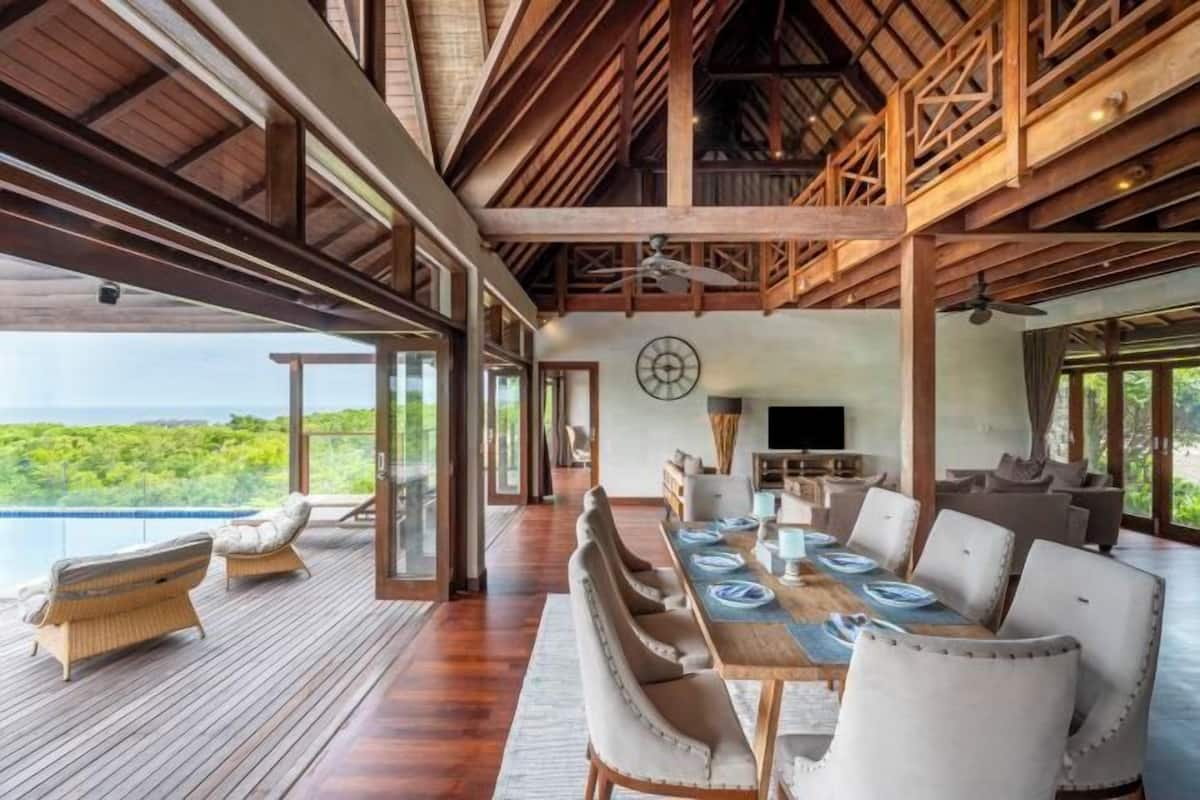 A spacious dining area with a long table set for six, a television, and a view of lush greenery.