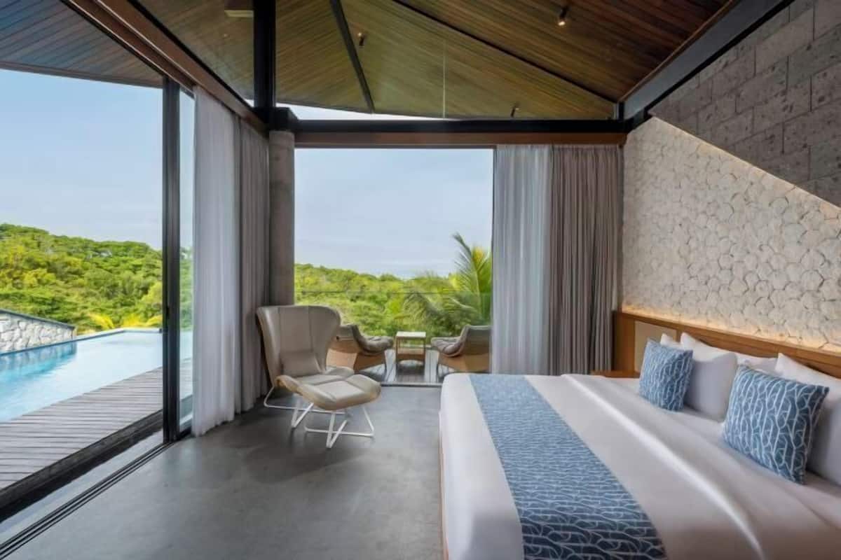 A modern hotel room with a large bed, a view of a swimming pool, and a balcony overlooking lush greenery.
