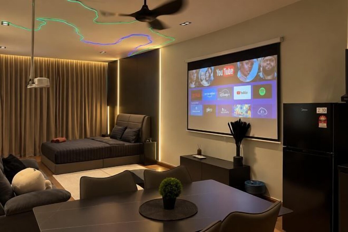 A modern living room with a bed, a dining table, and a projector screen displaying YouTube channels.