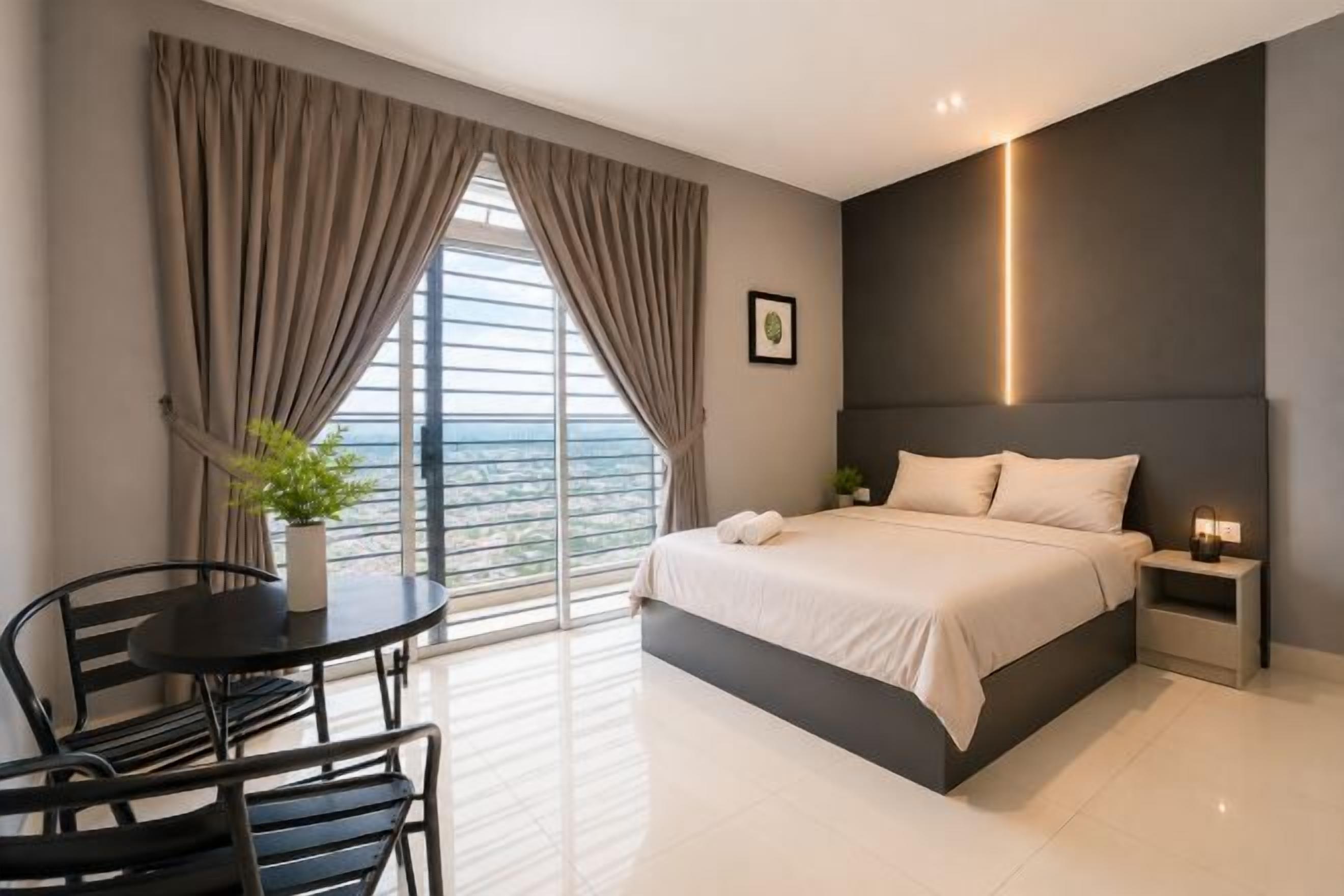 A modern hotel room with a large bed, a small table with chairs, a bedside table with a lamp, and a view through a large window.