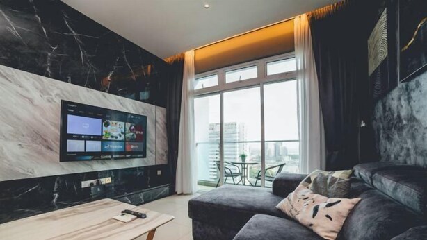 A modern living room with a large flat-screen TV mounted on the wall, a grey sofa with a decorative pillow, a small wooden table, and a large window with a view of the city. - Twin Galaxy Residences (Johor Bahru)
