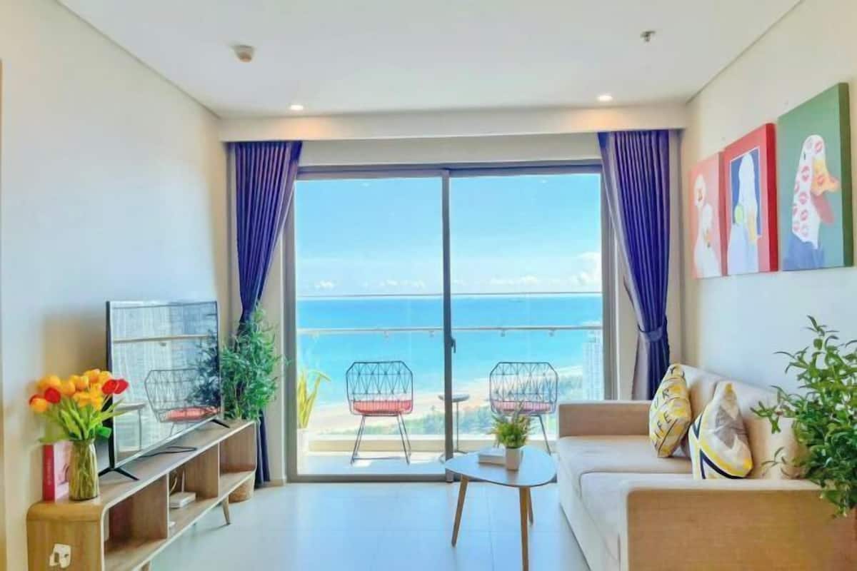 A modern living room with a sofa, two chairs, a TV, and a balcony with a view of the sea.