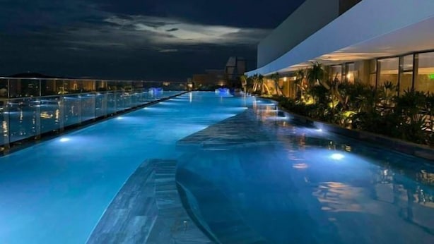 A nighttime swimming pool with a glass barrier, surrounded by a modern building with illuminated windows and palm trees. - The Song Vung Tau - Hong Hanh Apartments (Vung Tau)