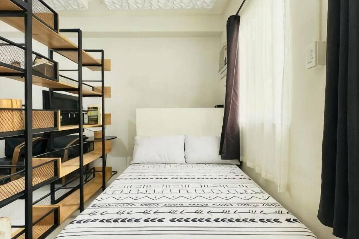 A narrow hotel room with a bed, a wooden bookshelf, and a window with curtains.