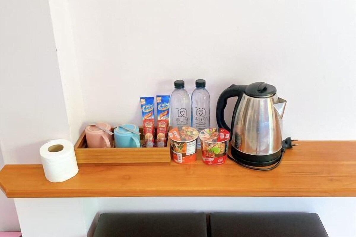 Une étagère en bois sur laquelle se trouvent un rouleau de papier toilette, deux paquets de mouchoirs, deux bouteilles d’eau, un thermos et une bouilloire électrique en acier inoxydable.