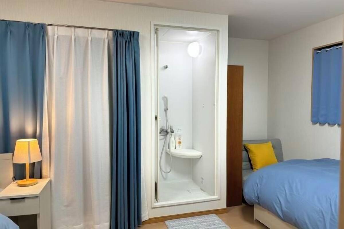 A hotel room with a shower, a bed with blue bedding, a nightstand with a lamp, and a blue curtain.