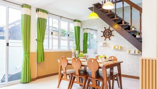 A dining area with a wooden table and chairs, a staircase with a ship's wheel, and large windows with green curtains. - Friend House BKK (Bangkok)