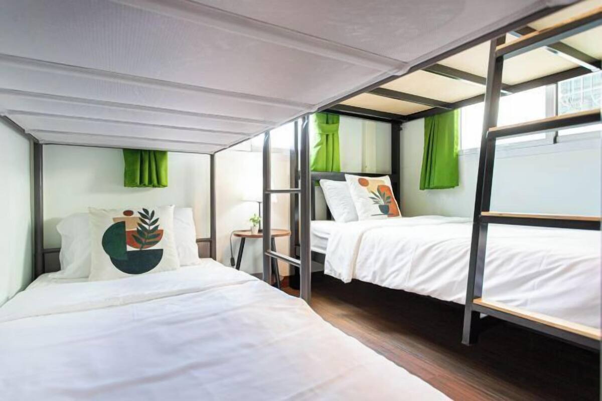 A modern, compact room with two beds and a small table, featuring green curtains and a minimalist design.
