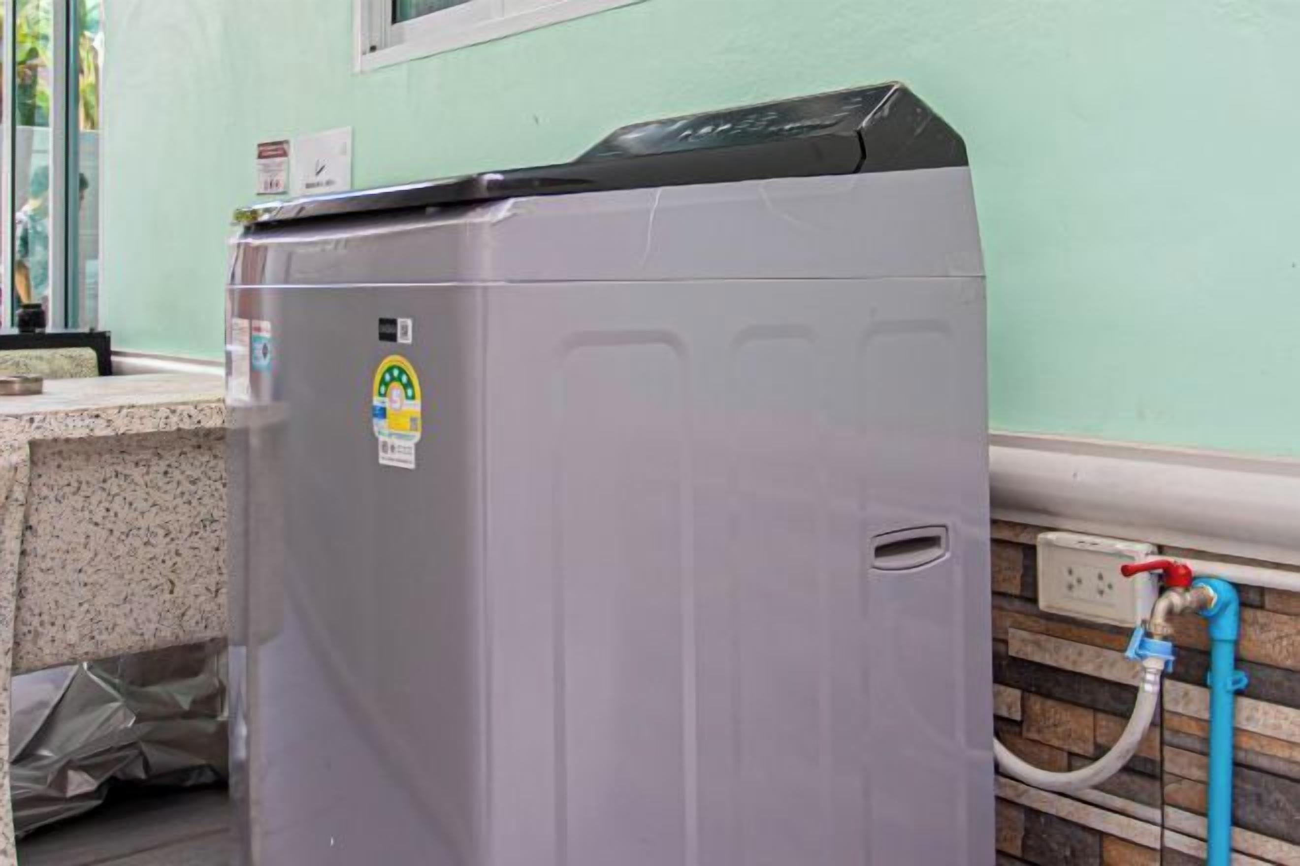 Een grijze wasmachine met een bedieningspaneel, een slang en een wandcontactdoos.