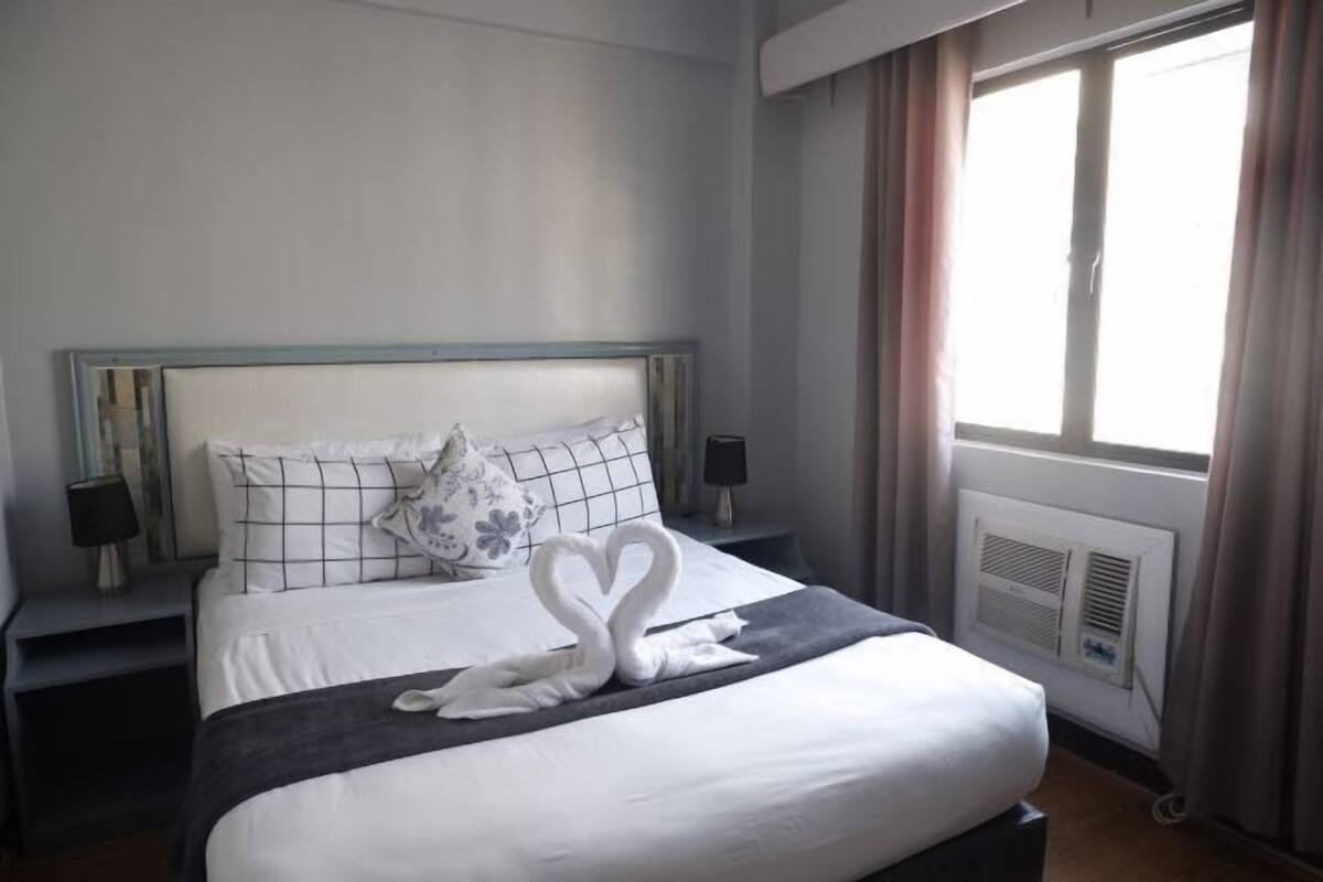 Un lit bien fait, agrémenté d’une serviette pliée en forme de cœur, une table de chevet avec une lampe et une fenêtre avec des rideaux.
