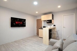 Un salon moderne avec un canapé gris, un téléviseur à écran plat fixé au mur, un coin cuisine équipé d’un four à micro-ondes et une porte donnant sur une autre pièce.