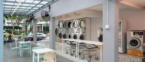 A laundry facility with multiple washing machines and dryers, a seating area with tables and chairs, and a glass ceiling.