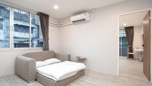 A modern hotel room with a bed, a small bedside table, a window with curtains, an air conditioning unit, and a view of the outside.