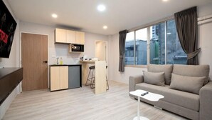 A modern living room with a sofa, a small coffee table, a kitchen area with a microwave, and a large window with curtains.