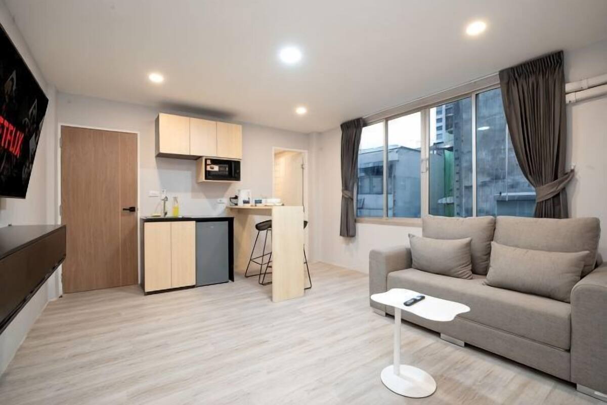 A modern living room with a sofa, a small coffee table, a kitchen area with a microwave, and a large window with curtains.