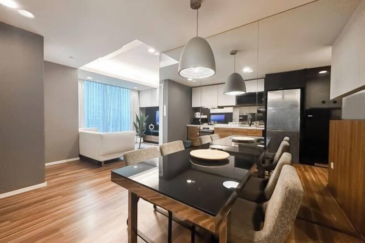 Un espace de salle à manger moderne avec une table noire, plusieurs chaises, des luminaires suspendus et une cuisine en arrière-plan.