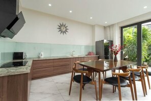 A modern kitchen with a dining table and chairs, a TV mounted on the wall, and a large window providing a view of greenery.