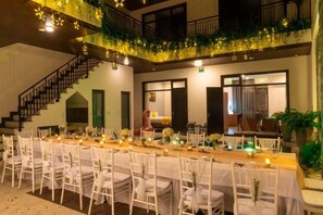 An indoor banquet hall with a long table set for dining, surrounded by white chairs, and a staircase leading to an upper level.