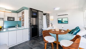 A modern kitchen with white cabinets, a microwave, and a sink. There's a dining area with a round wooden table and two chairs. A sofa with cushions is against the wall, and a TV is mounted above it.