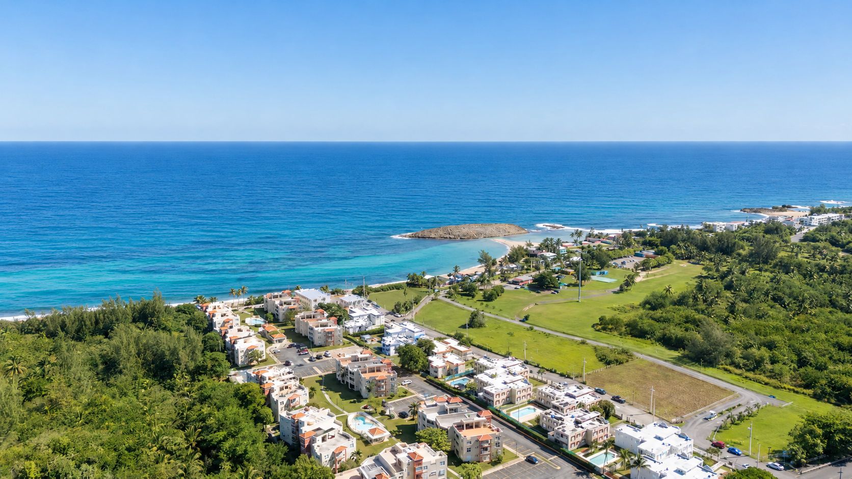 Una zona residenziale lungo la costa, con edifici, un mare blu cristallino e un piccolo promontorio roccioso.