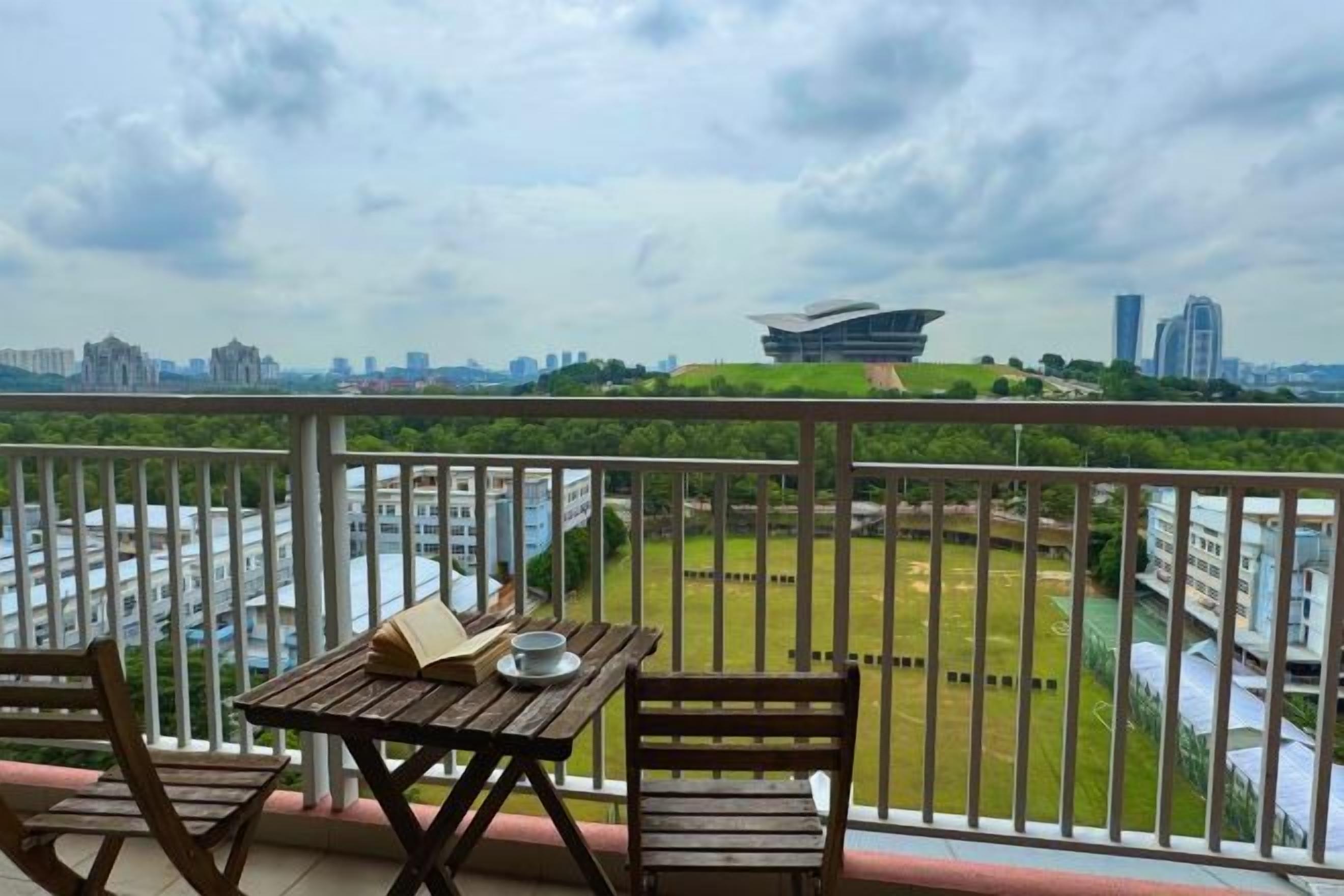 A balcony with a table and chairs, overlooking a cityscape with buildings and a sports field.