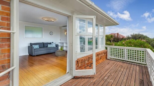 Une porte ouverte donne sur une pièce où se trouvent un canapé et une petite table, ainsi qu’une terrasse en bois et une clôture blanche.