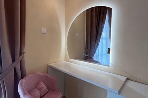 Un dormitorio con una tocador curvo, una silla rosa y un ventanal con cortinas.