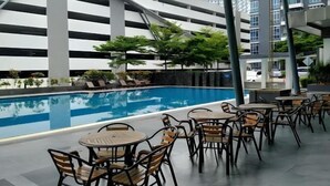 Un área de piscina en la azotea con sillones y mesas, rodeada por un edificio moderno y un aparcamiento.