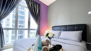 Habitación de hotel con cama, mesita con toallas y un secador, florero con flores y vistas a edificios a través de amplias ventanas.