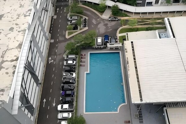Una piscina rodeada por un estacionamiento con coches, un edificio de techo plano y una carretera con flechas direccionales.