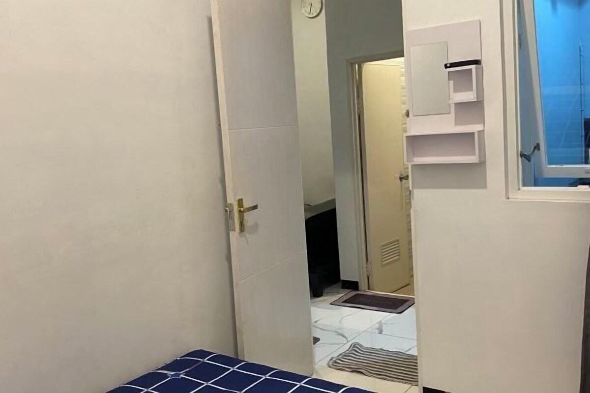 Passadís estret amb una porta que dona a una habitació amb un llit, una finestra petita i una estanteria penjada a la paret.