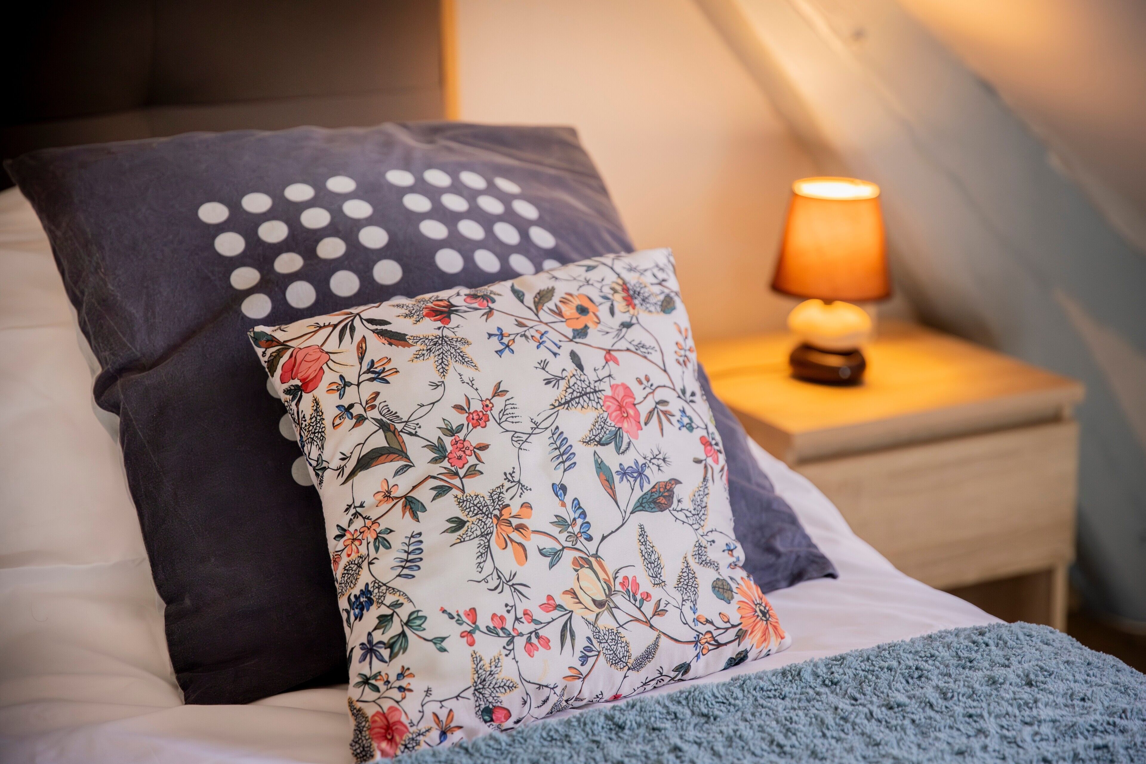A bed with a floral pillow, a polka-dotted pillow, and a nightstand with a lamp.