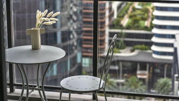 A balcony with a table and a chair, a vase with white flowers, and a cityscape view through large windows. - Waterview 1Bedroom Plus Study Room at Wentworth Point (Wentworth Point)