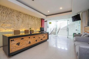 A reception area with a wooden reception desk, a wall-mounted TV, and a staircase in the background. - RedDoorz @ First Homestay Bypass Kuta (Bali)