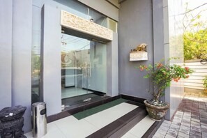 A modern building entrance with a glass door, a potted plant, and a small trash bin. - RedDoorz @ First Homestay Bypass Kuta (Bali)