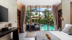 A bedroom with a bed, a desk, a TV, and a view of a swimming pool and tropical plants. - Villa Na (Bali)