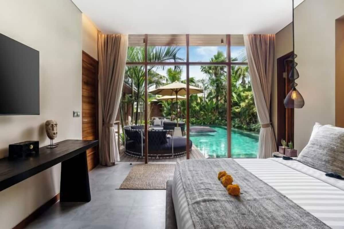 A bedroom with a bed, a desk, a TV, and a view of a swimming pool and tropical plants.