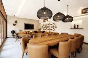 A modern dining area with a wooden table and leather chairs, pendant lighting, and a wall-mounted TV. - Villa Na (Bali)