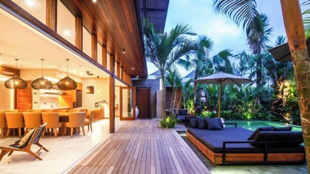 A modern outdoor patio with wooden flooring, a dining area with orange chairs, a poolside seating area, and lush tropical plants. - Villa Na (Bali)