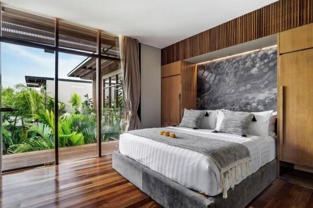 A modern bedroom with a large bed, wooden headboard, and a view of lush greenery.