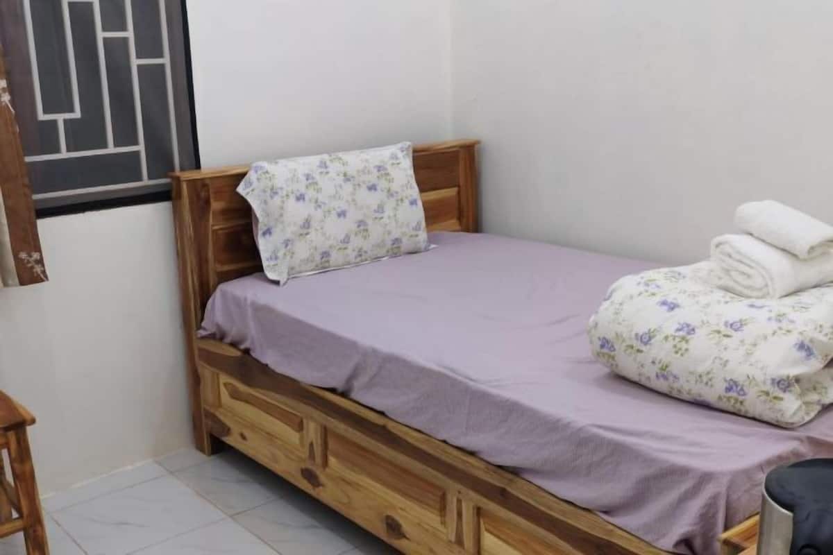 A single bed with a wooden frame, a purple bedspread, and a floral pillow. There is a wooden nightstand beside the bed with a folded towel on it.