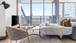 A modern bedroom with a bed, a chair, a desk, a TV, and a large window with a city view. - A Stylish 3BR Apt with Harbour Views, FREE Parking (Melbourne)