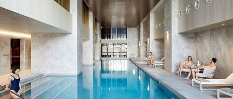 An indoor swimming pool with a long, narrow design, a person cleaning the pool, and lounge chairs along the sides.