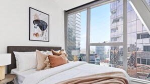 A modern hotel room with a large window, a neatly made bed, a framed picture on the wall, and a city view. - A Stylish 3BR Apt with Harbour Views, FREE Parking (Melbourne)