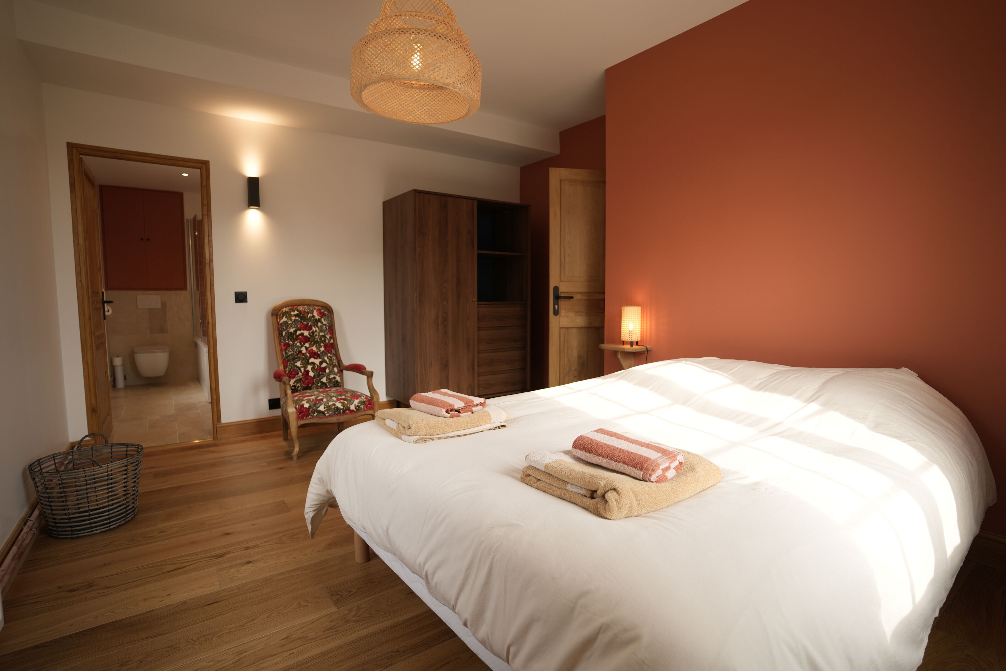 Une chambre avec un grand lit, un plancher en bois, une lampe en osier et un mur rouge.