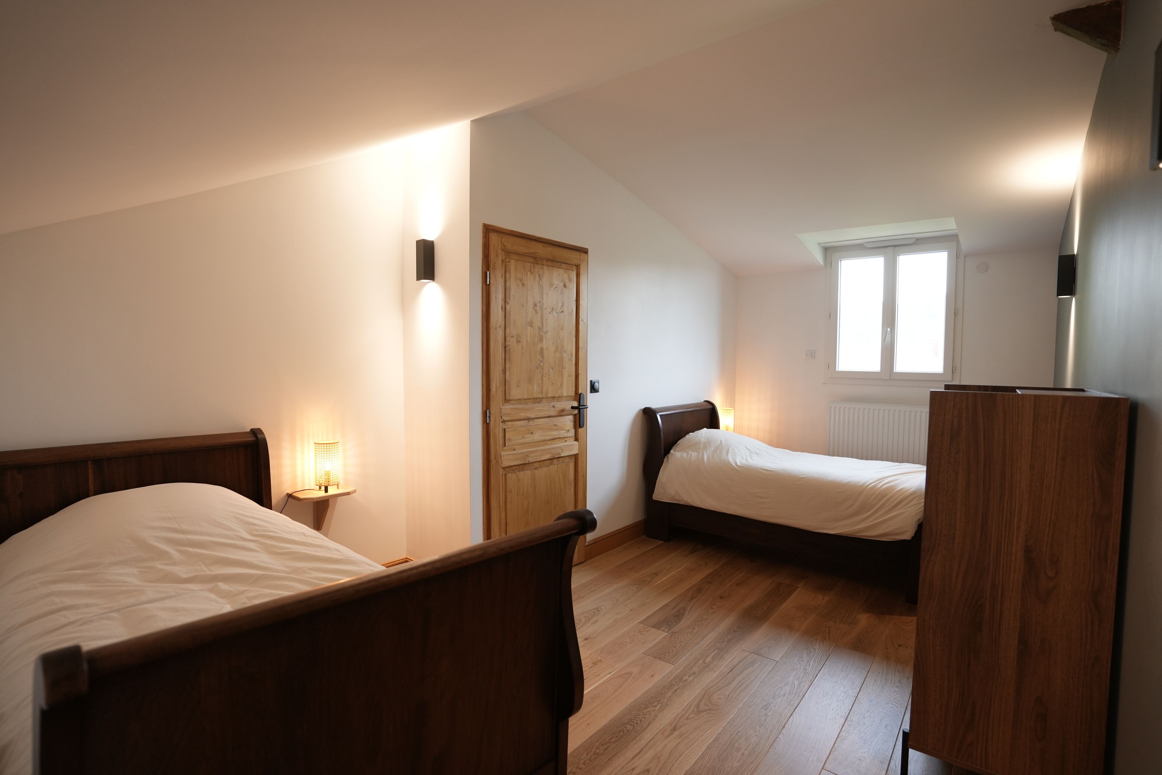 Quarto com duas camas de madeira, um guarda-roupa de madeira, uma mesa de cabeceira com abajur e uma janela com persianas.