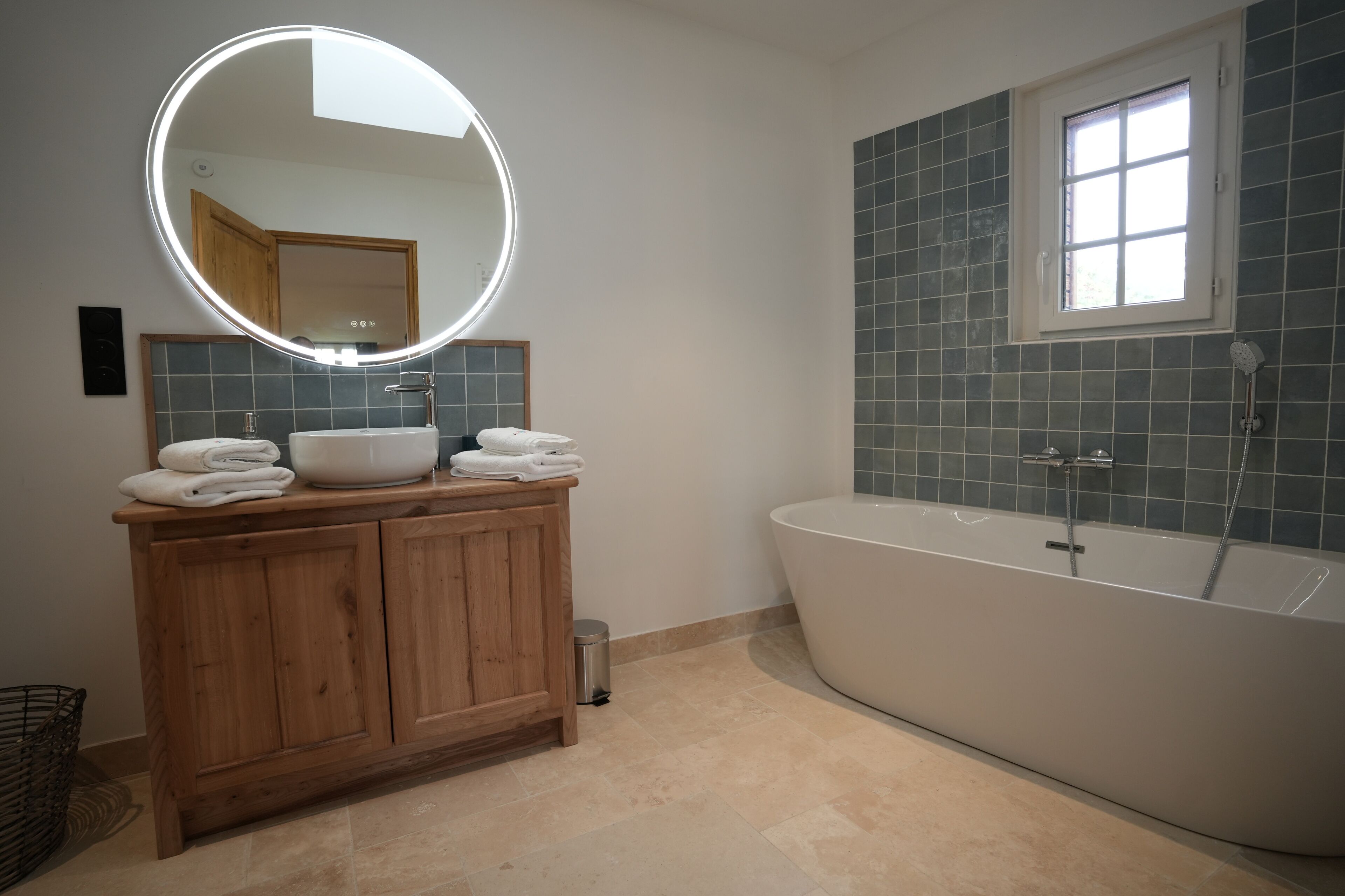 A modern bathroom with a wooden vanity, a round mirror with lights, a freestanding bathtub, and a window.